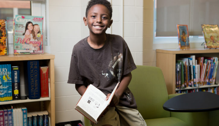 Black male middle-school student with book sitting on arm of chair and smiling for literacy program article