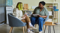 Black female social worker talking to teenage girl while they sitting on armchairs at office for article on mental health supports