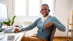 Black woman in pale blue sweater wearing wired headphones in front of a computer on a desk and smiling for article on microcredentials for educators.