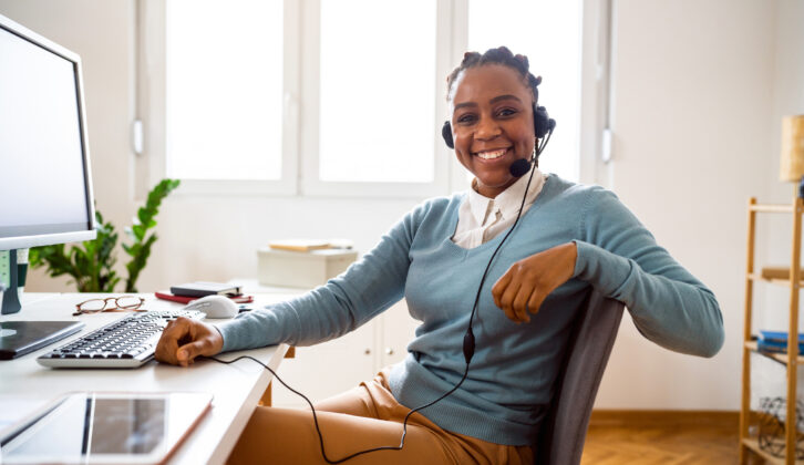 Black woman in pale blue sweater wearing wired headphones in front of a computer on a desk and smiling for article on microcredentials for educators.