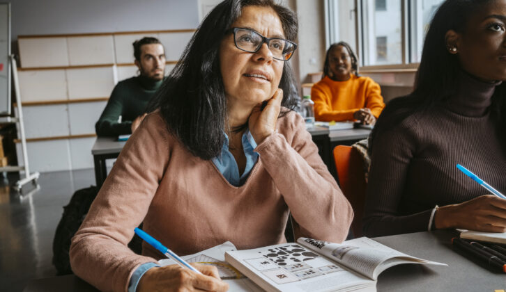Older woman in glasses at classroom desk with book open, looking toward front of classroom for article on prior learning