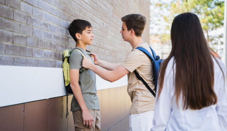 1 school boy pushing another against a wall, holding him by backpack straps while a school girl watches for article on trauma-informed approach