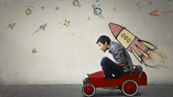 Young Asian male squished into a red toy car in front of a gray wall with spaceship and planets, for article on student momentum