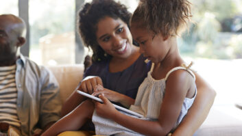 Black daughter sitting on Black mother's lap. Child has tablet, and mom is pointing something out. For article on successful literacy program