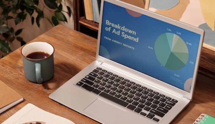 laptop on a wooden desk. The open laptop has a pie chart and reads Breakdown of Ad Spend