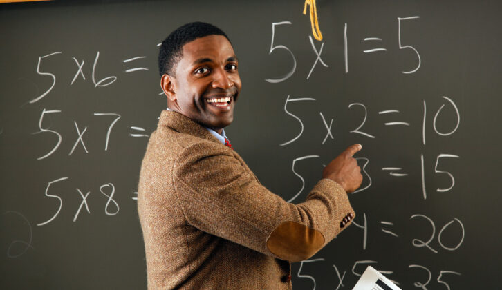 Black male teacher in sportscoat pointing at chalkboard with math equations on it, smiling