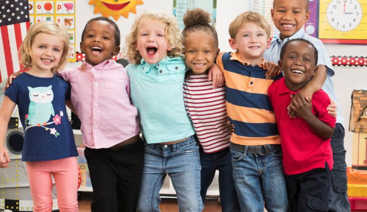 seven smiling preschoolers standing in a row in a classroom with arms around each other's shoulders.