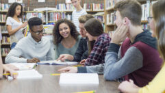 A multi-ethnic group of high-school students seated around a library, smiling, with one pointing at something in a book.