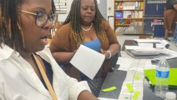 2 Black female educators sitting at a long table covered in papers going over work together.