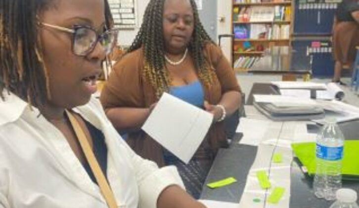 2 Black female educators sitting at a long table covered in papers going over work together.