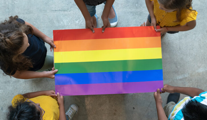 Children standing in a circle holding the edges of a flat rainbow flag