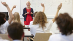Female teacher and class with hands raised for article on teacher authority